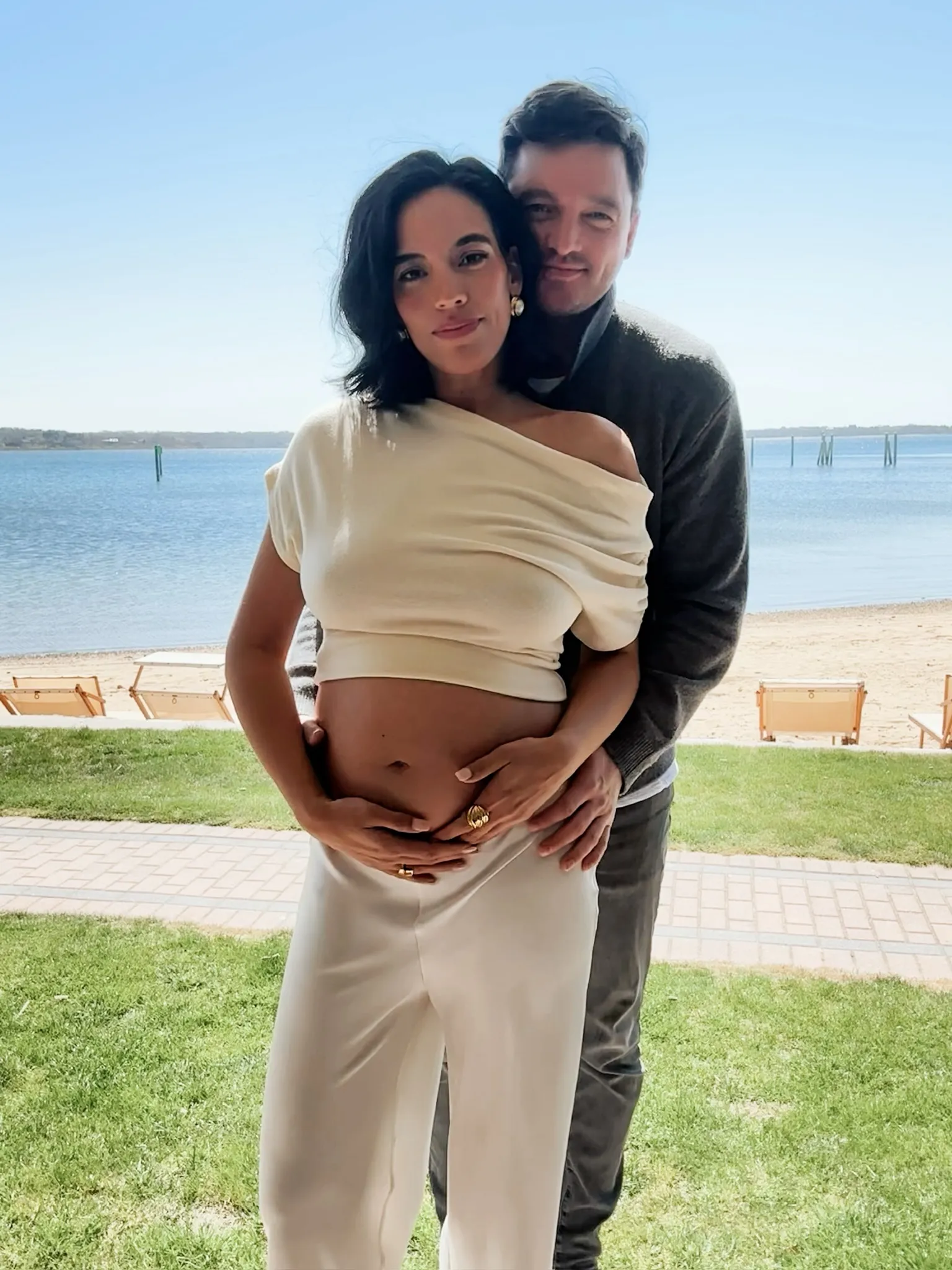 A man stands behind a pregnant woman, holding her belly, with a beach and water in the background.
