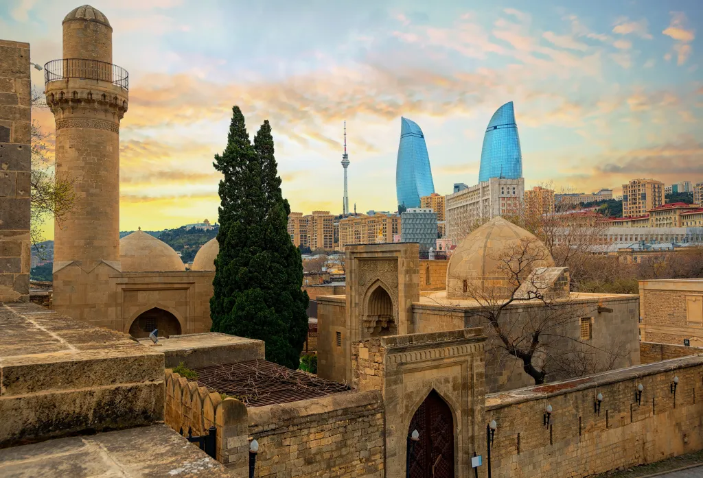 Old mosques and the Shirvanshahs Palace in Baku's Old Town, with the Flame Towers and TV tower in the background at sunset.