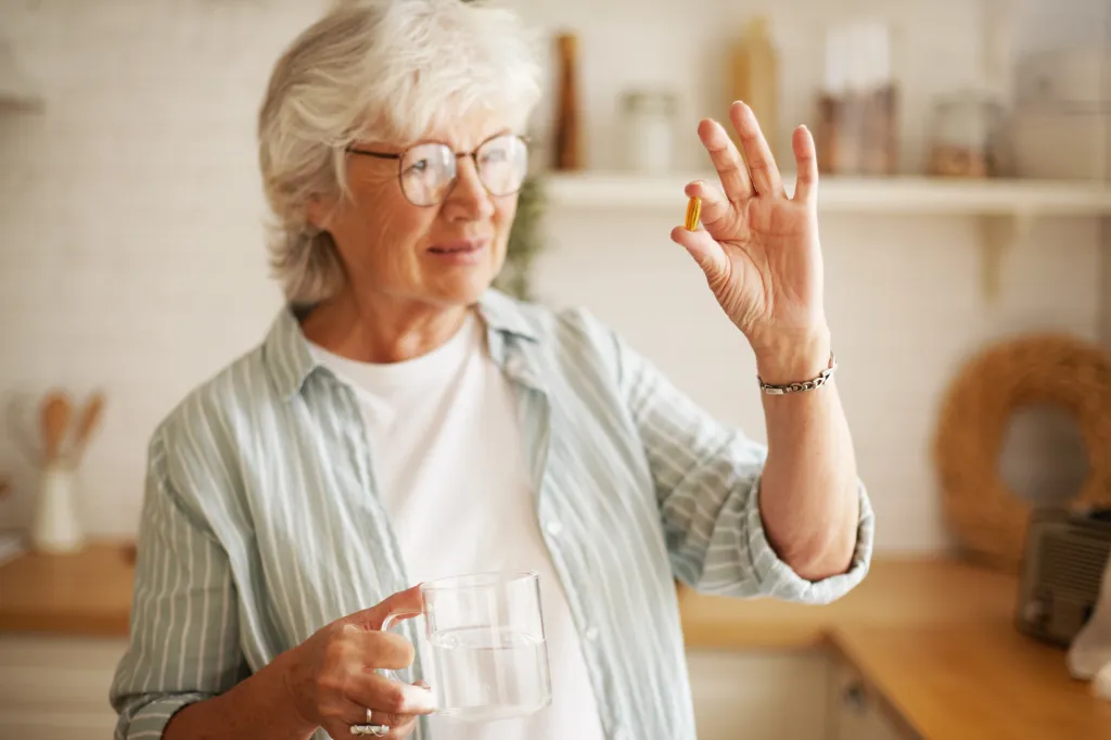 A sixty-year-old woman in glasses holds an omega-3 capsule and a glass of water.