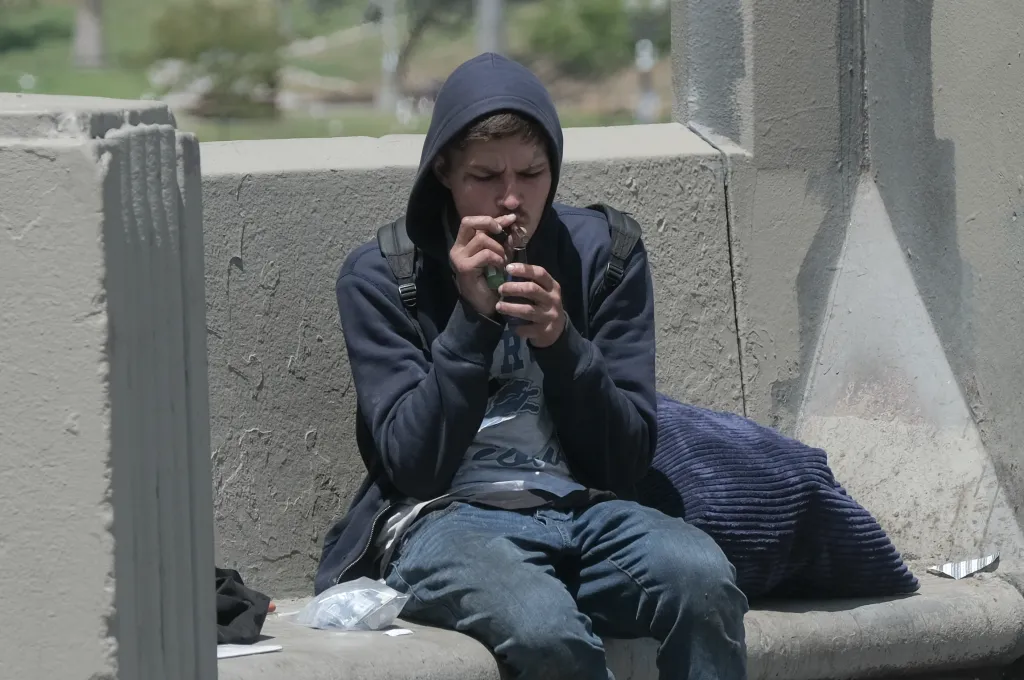 A young man in a hooded sweatshirt lights a pipe at a bus stop.