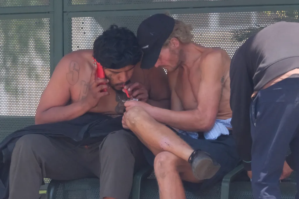 Two men engaged in drug use at a bus stop, with one talking on a red phone.