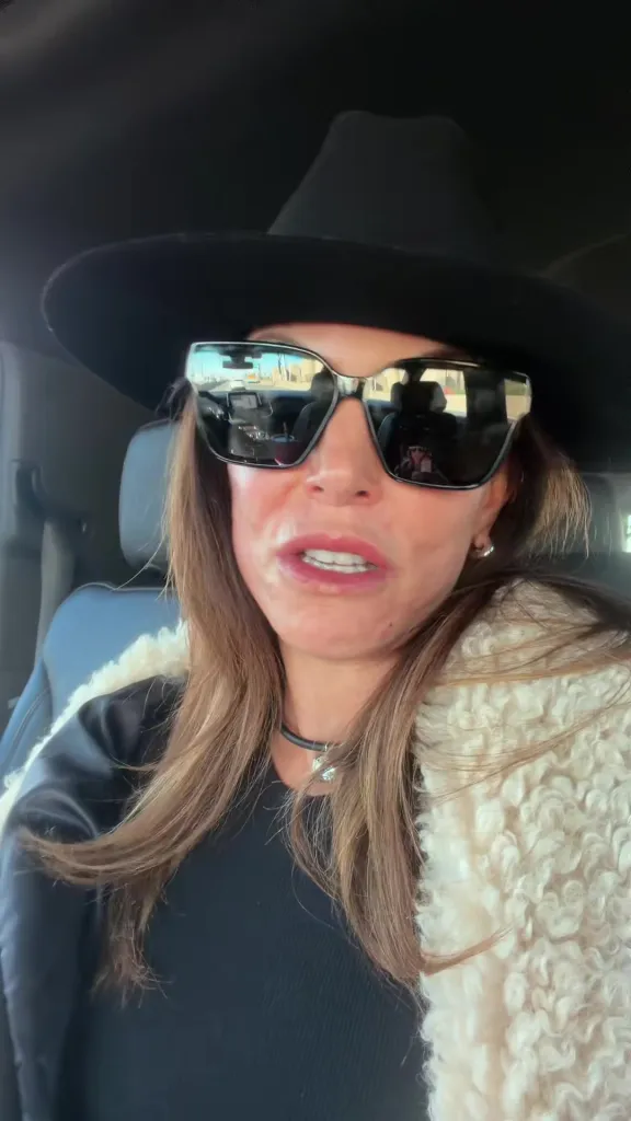Bethenny Frankel, wearing a black hat, black sunglasses, and a cream fluffy coat, looking into the camera.