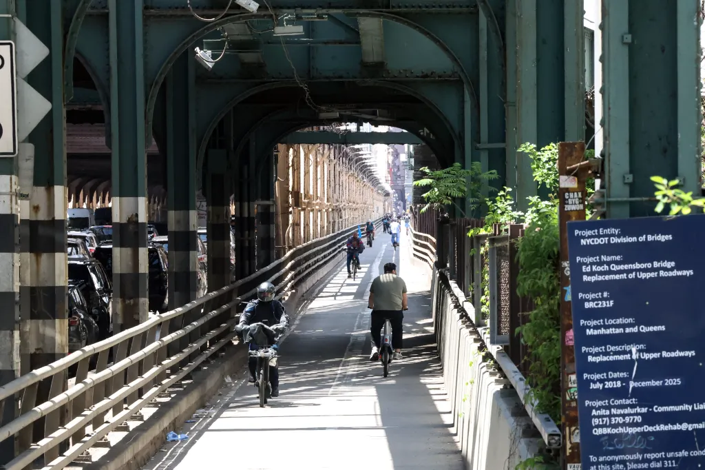 Cyclists riding on the Queensboro Bridge bike path, with traffic visible in the distance and a sign detailing bridge repairs.