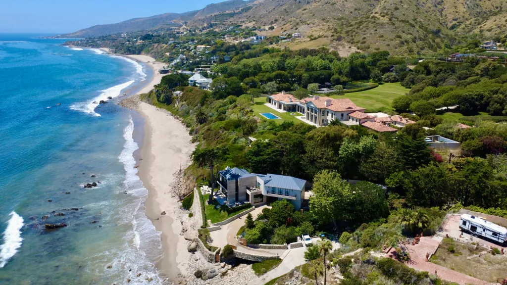 Aerial view of the Malibu coast showing Daniel Nadler's mansion with a pool, and other houses along the shoreline with mountains in the background.