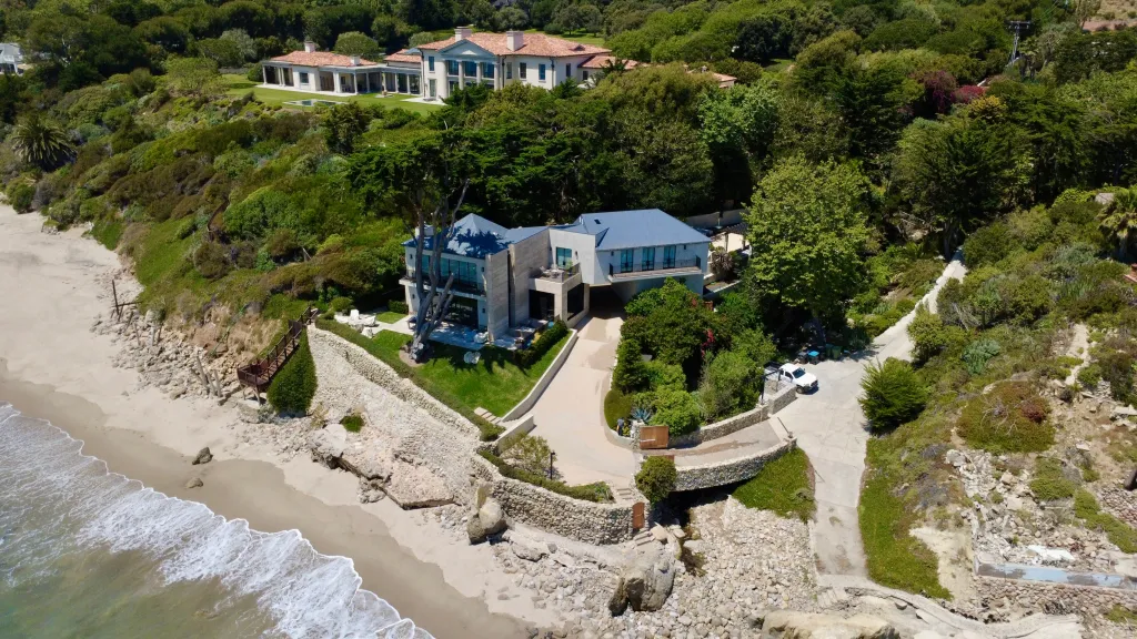 Aerial view of a coastal property featuring a mansion at the top of a green hill, and a smaller, modern house with a blue roof closer to a sandy beach.