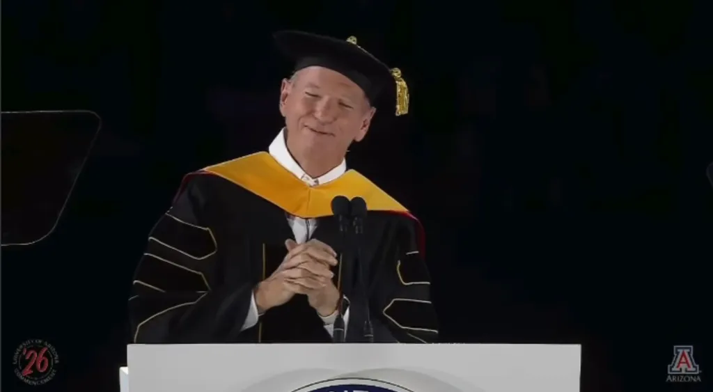 Eric Schmidt at a podium, wearing a graduation cap and gown, appearing to laugh or smile while being booed during his commencement speech.
