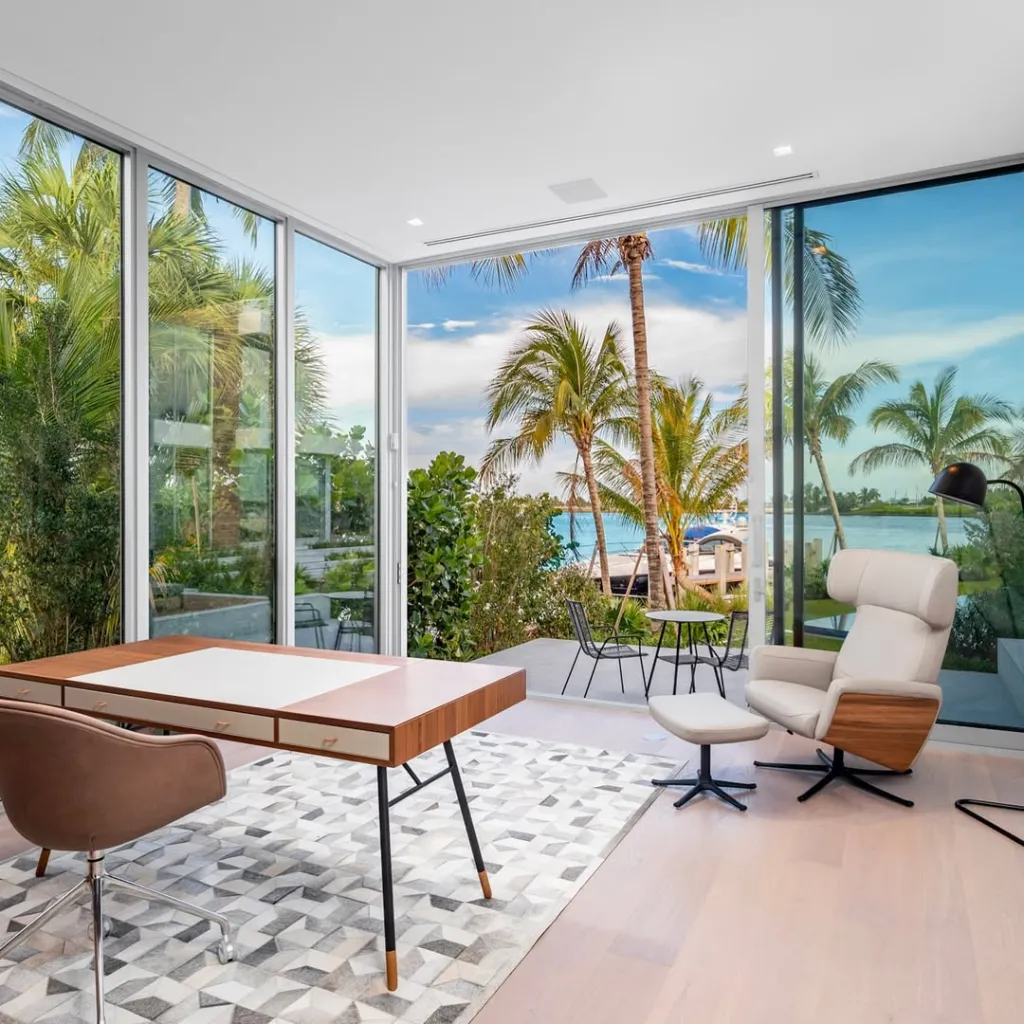 Office with a wooden desk, office chair, a cream armchair, and an ottoman, all facing a wall of windows overlooking a tropical landscape with palm trees and a body of water.