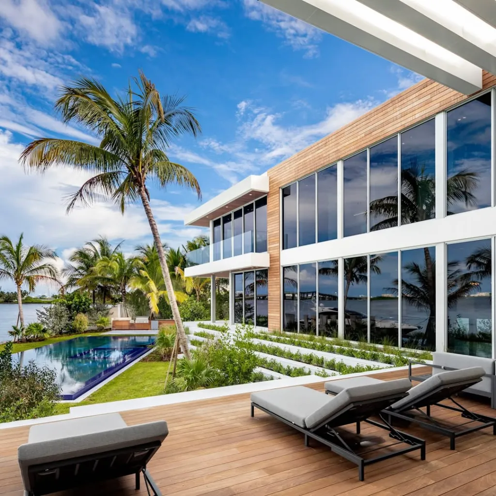 Modern mansion with a wooden deck, lounge chairs, infinity pool, and palm trees overlooking the water.