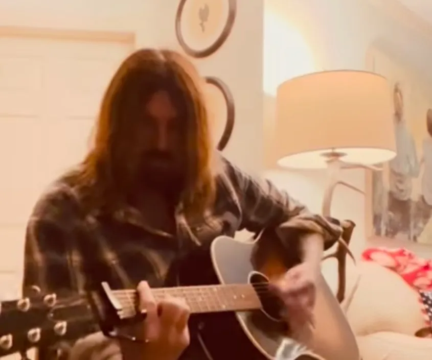 Billy Ray Cyrus singing a song and playing an acoustic guitar.