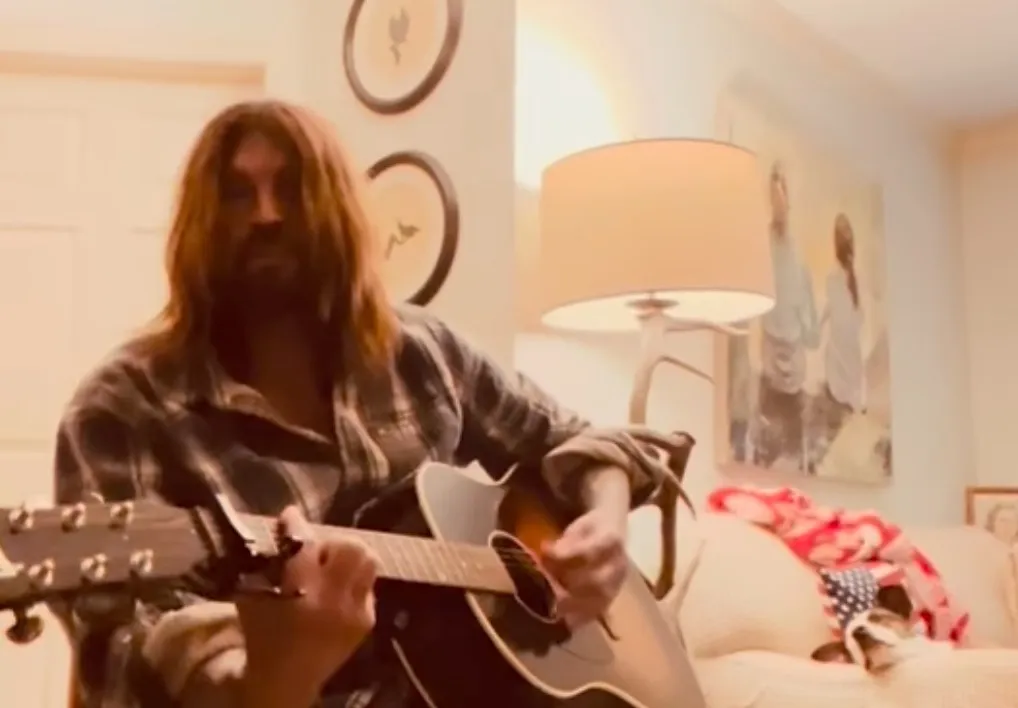 Billy Ray Cyrus playing an acoustic guitar indoors.