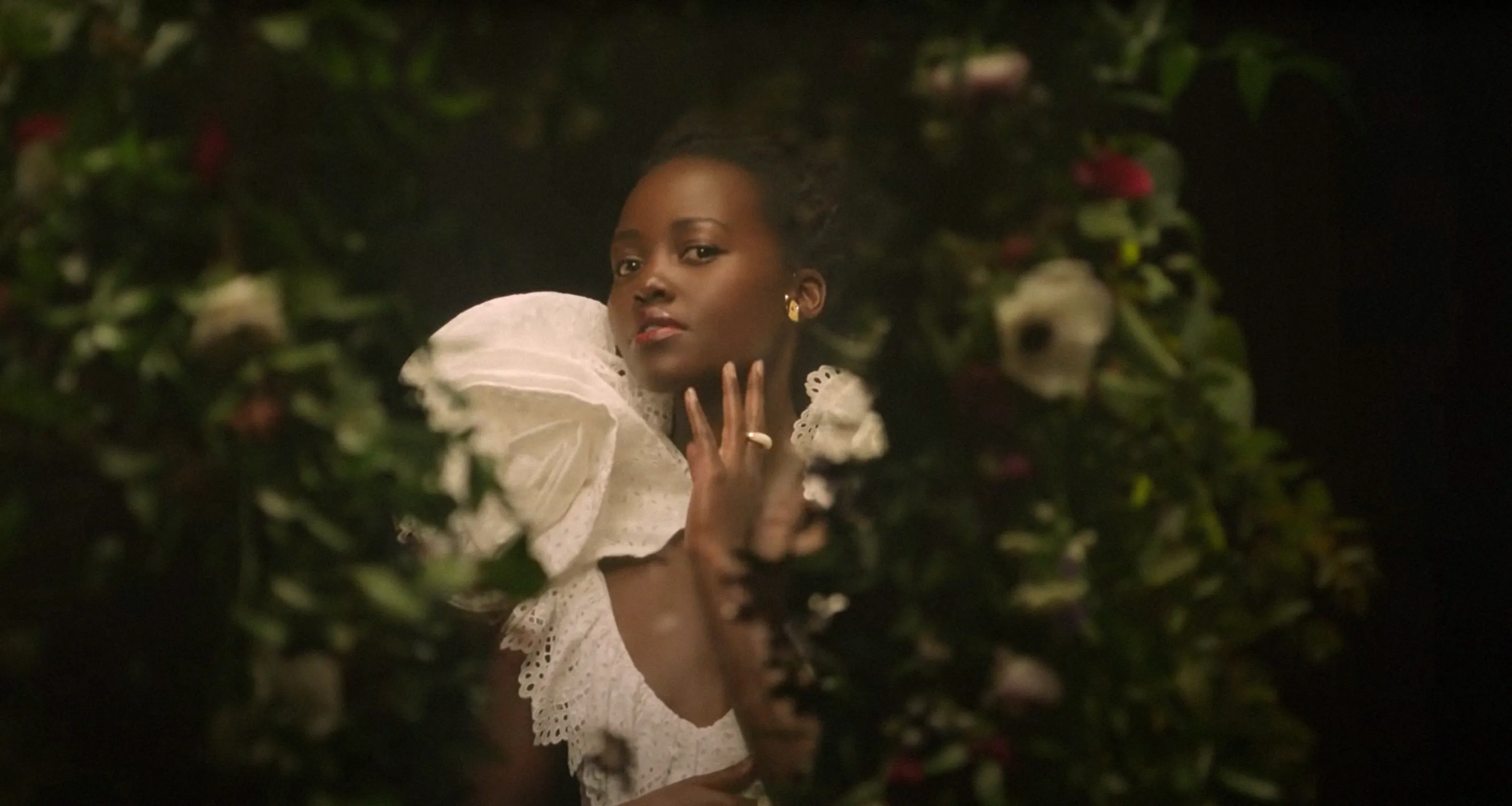 Lupita Nyong'o, in character from the movie Black Is King, poses in a white ruffled top surrounded by greenery.