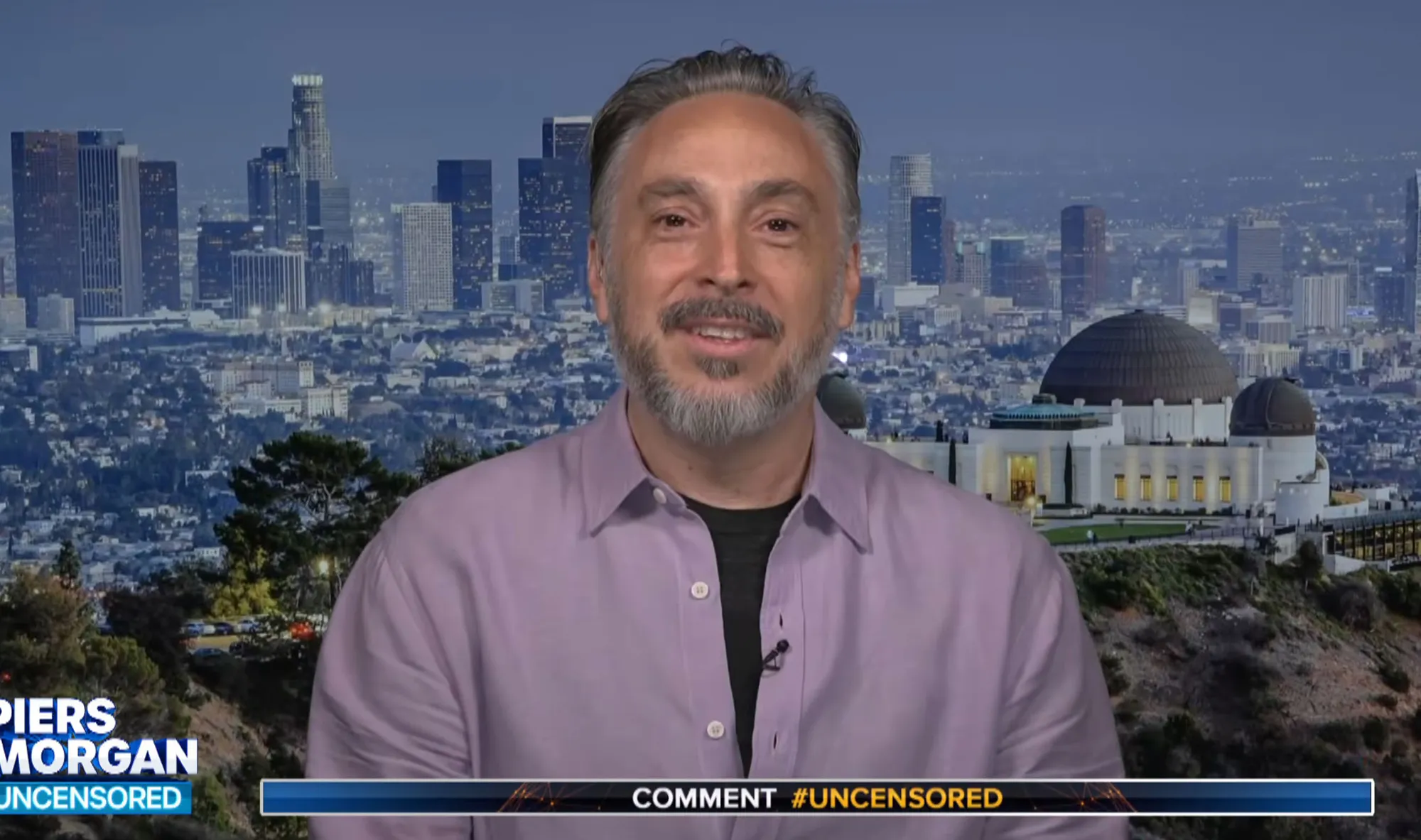 A man with grey hair and beard wearing a lavender shirt smiles in front of a cityscape at dusk, with the Griffith Observatory visible on the right.