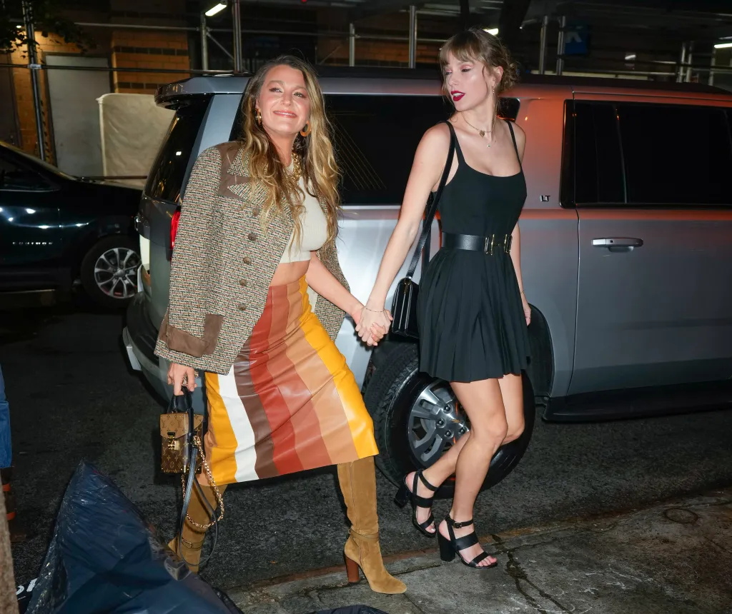 Blake Lively, in a striped skirt, brown boots, beige crop top and brown blazer, and Taylor Swift , in a black mini dress and black heels, holding hands in New York City.