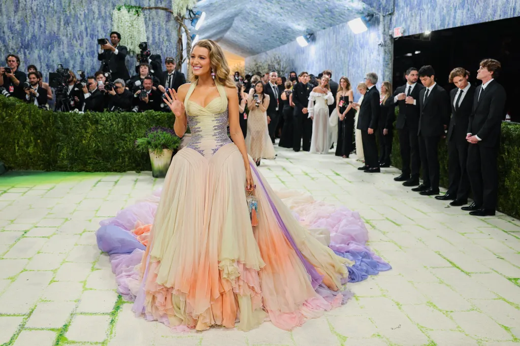 Blake Lively poses at the Met Gala in a long, flowing dress that transitions from pale yellow to orange, pink, and purple.