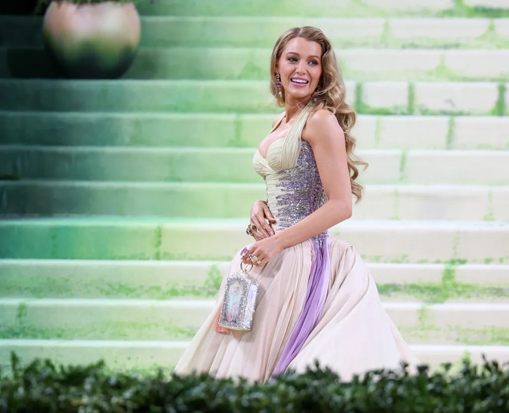 Blake Lively in a sparkling bodice and flowing cream and lavender skirt holds a jeweled purse while standing on green steps.