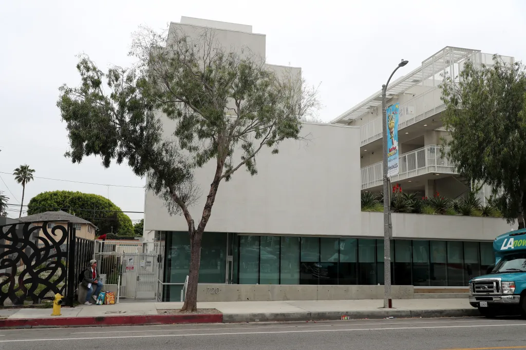 Street view of an apartment building with a woman sitting on the sidewalk and a turquoise 