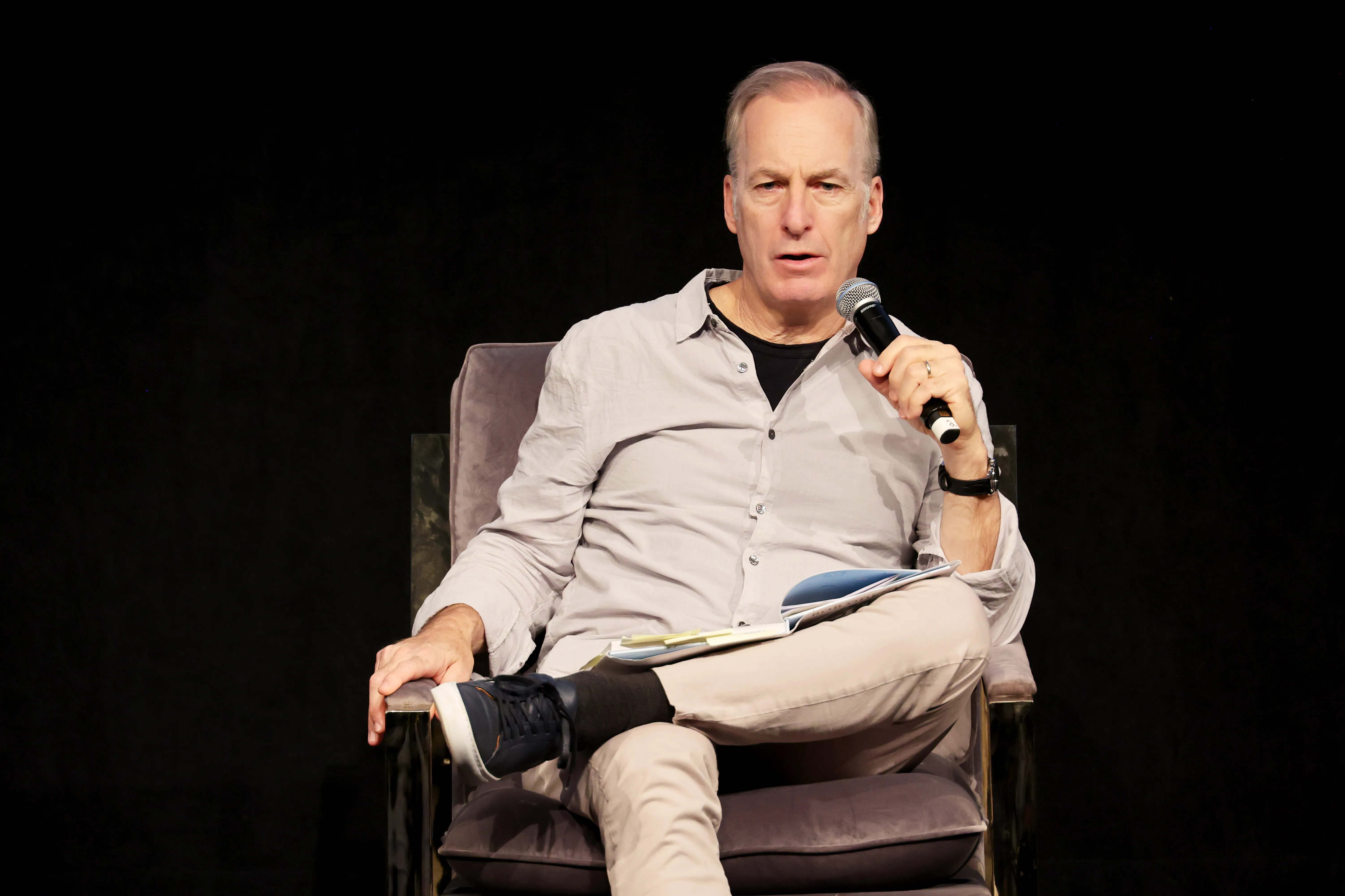 Bob Odenkirk speaking onstage while holding a microphone and a book during a book signing.