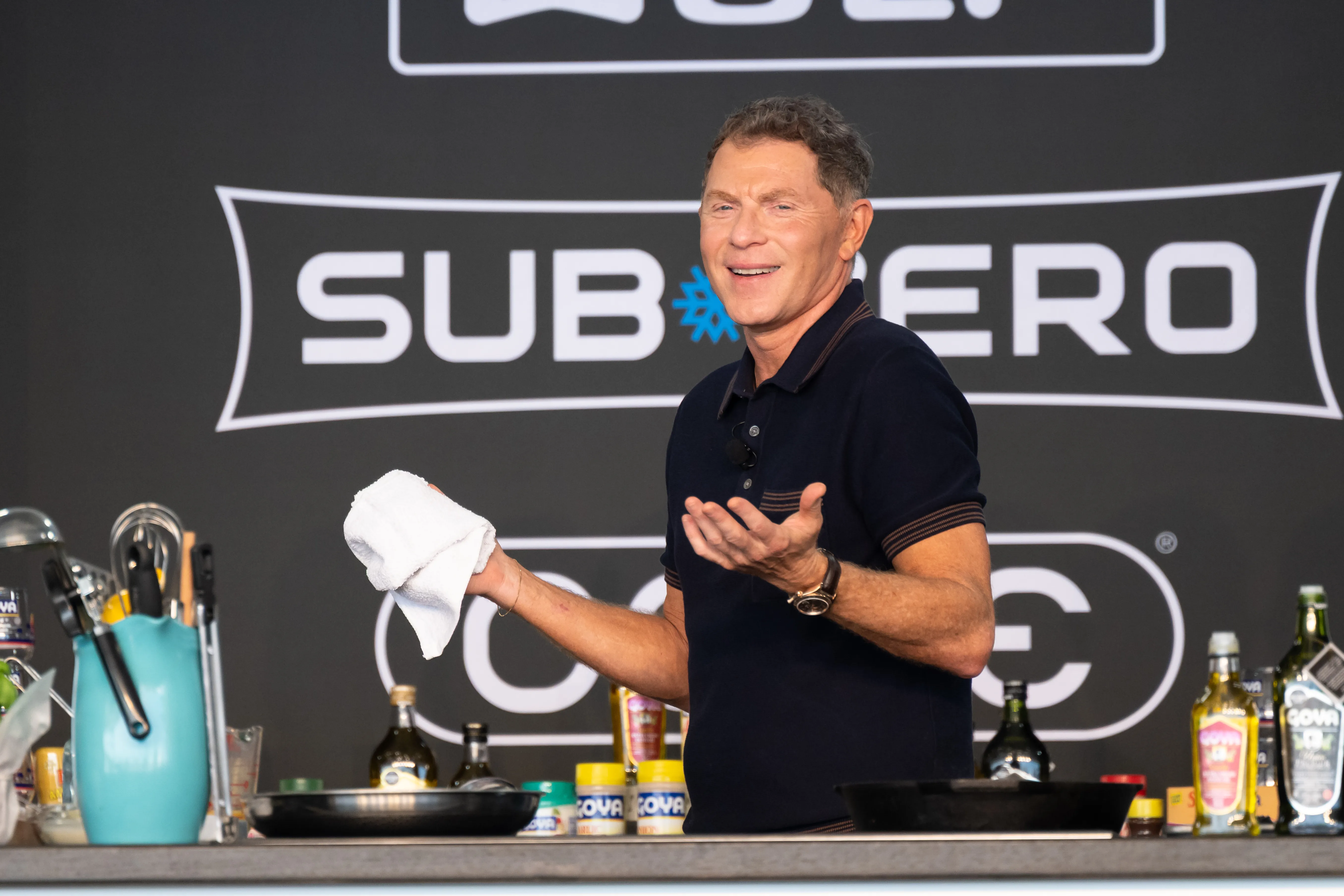 Bobby Flay on stage during a cooking demonstration, holding a white towel in one hand and gesturing with the other.