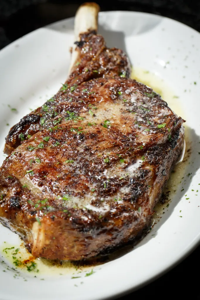 A bone-in ribeye steak on a white plate with butter and herbs.