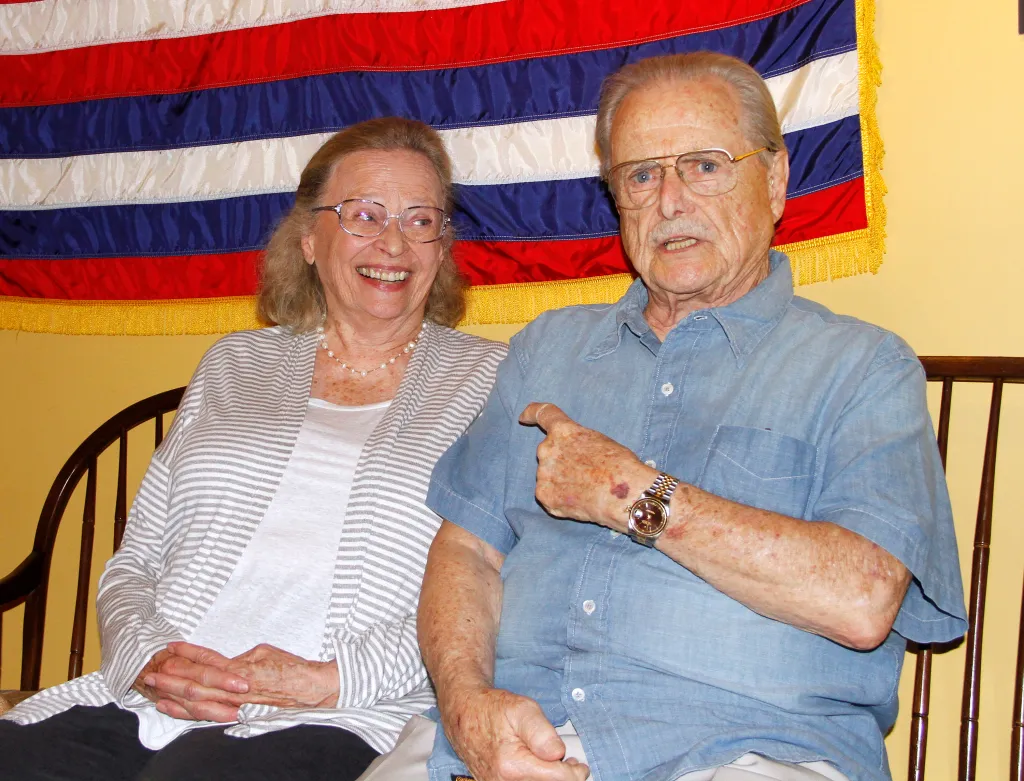 Actors Bonnie Bartlett and William Daniels sitting with a flag behind them.