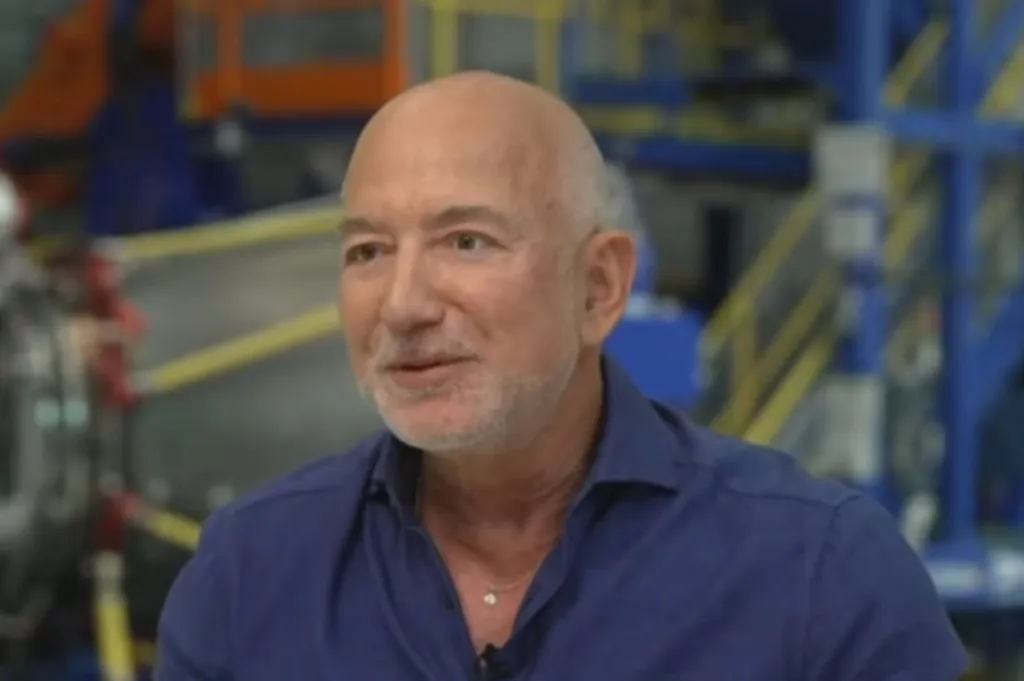Jeff Bezos in a blue shirt with a small diamond necklace, speaking.