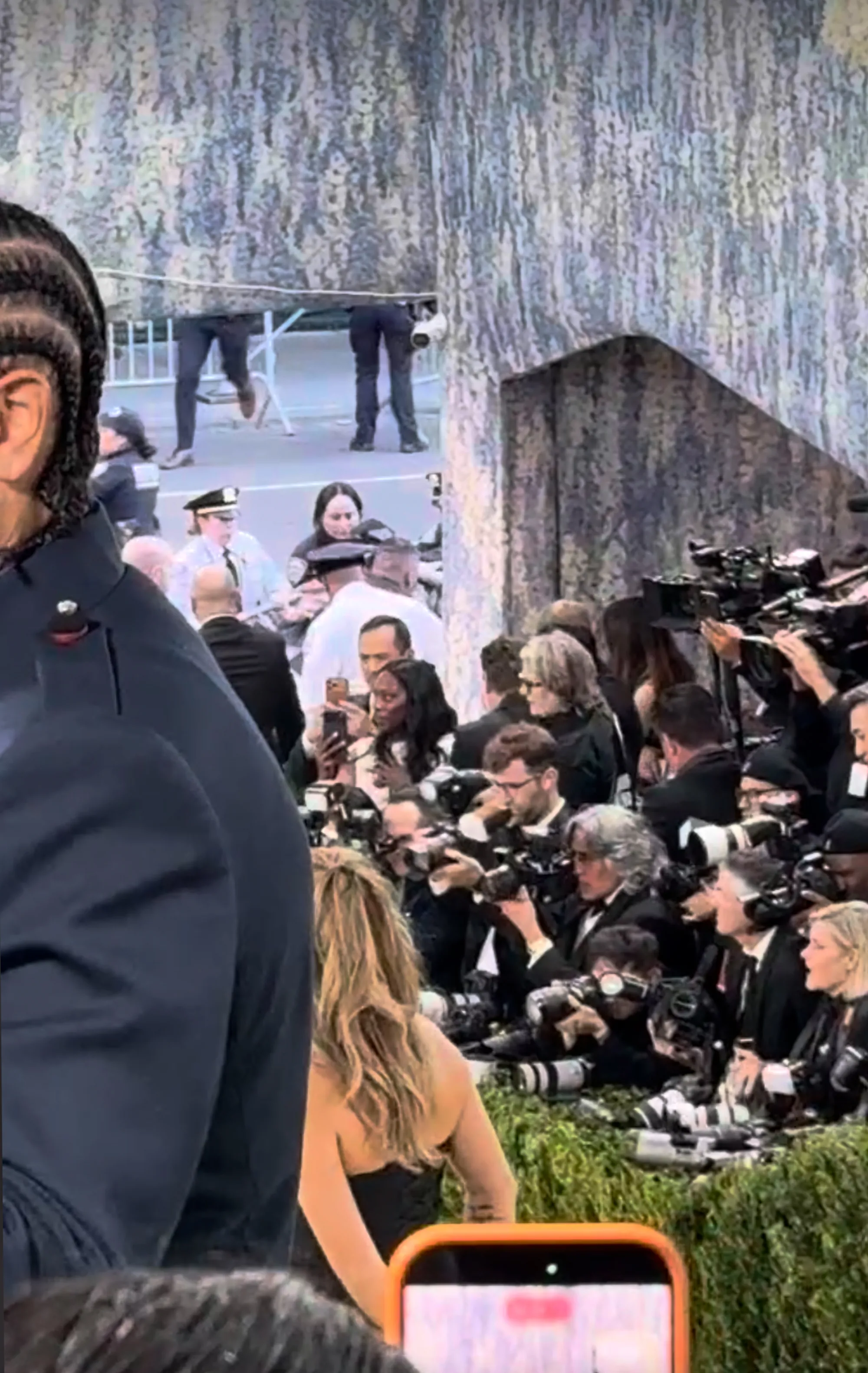 A man with braids and a dark coat on the left, an anti-Bezos protester breaking through a red carpet railing at the Met Gala 2026, surrounded by police and photographers.