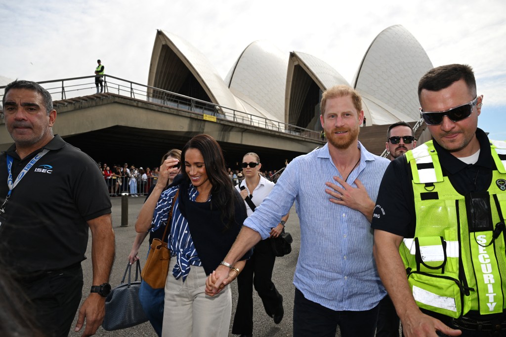 Prince Harry, in a blue shirt and dark pants, and Meghan Markle , in a blue and white striped shirt and beige pants, walk surrounded by security with the Sydney Opera House in the background.