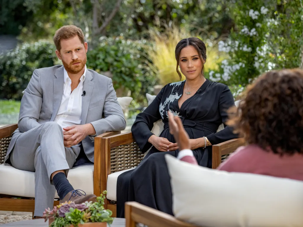 Meghan Markle and Prince Harry seated in chairs outdoors, with Oprah Winfrey's back to the camera, during an interview.