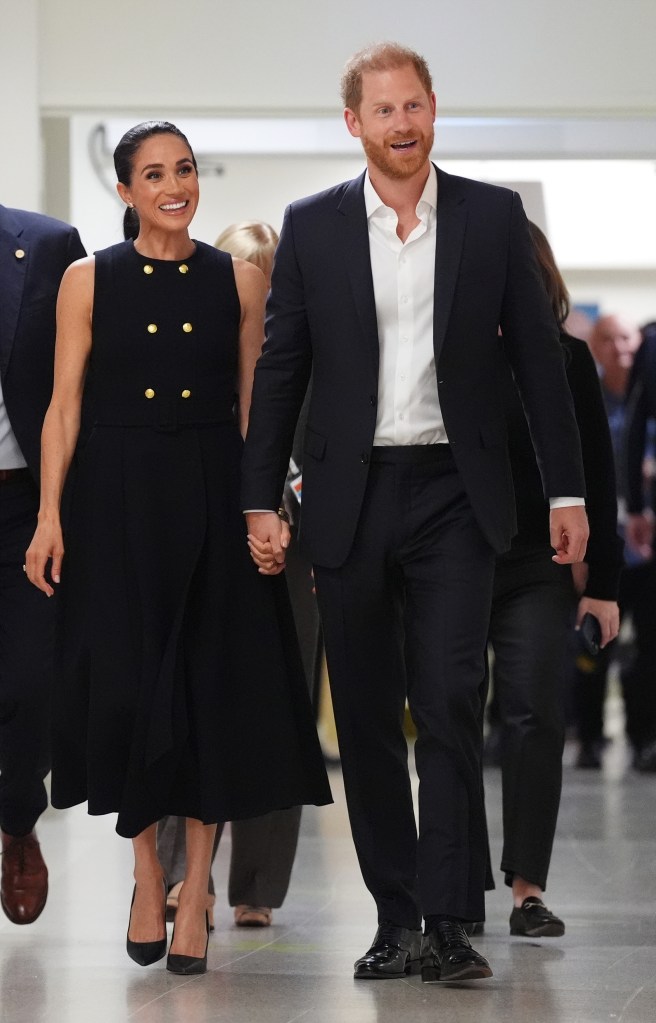 Prince Harry, in a black suit and white shirt, and Meghan Markle, in a sleeveless black dress with gold buttons, holding hands and smiling while walking.