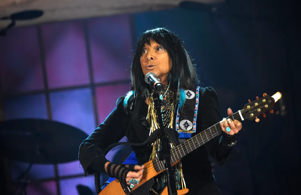 Buffy Sainte-Marie performs on stage, playing a guitar and singing into a microphone.