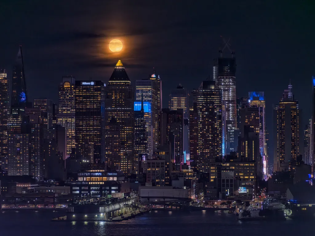 The Super Blue Moon rises over the Manhattan cityscape at night, reflecting on the water in the foreground.