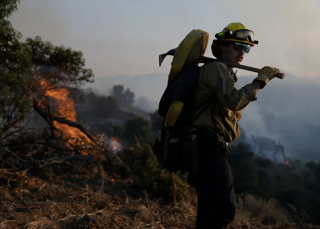 A Cal Fire firefighter with a tool and hose over their shoulder stands in front of a brush fire in Simi Valley, California.