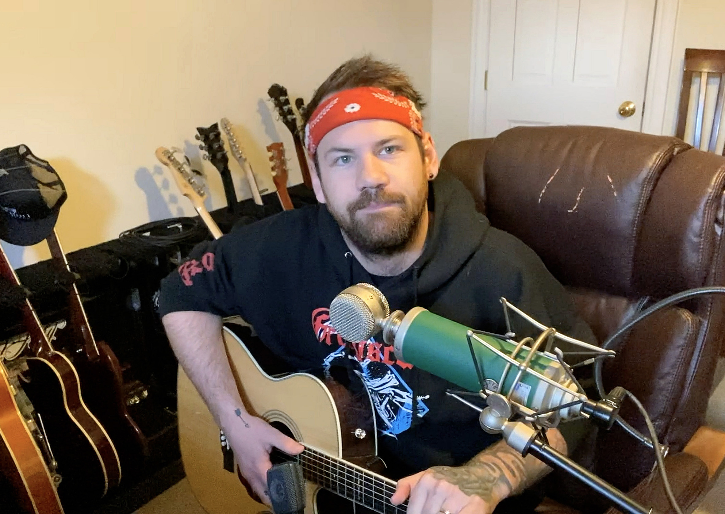 Caleb Shomo of Beartooth performs during a virtual festival, playing an acoustic guitar while sitting in a brown leather chair, with several guitars visible in the background.