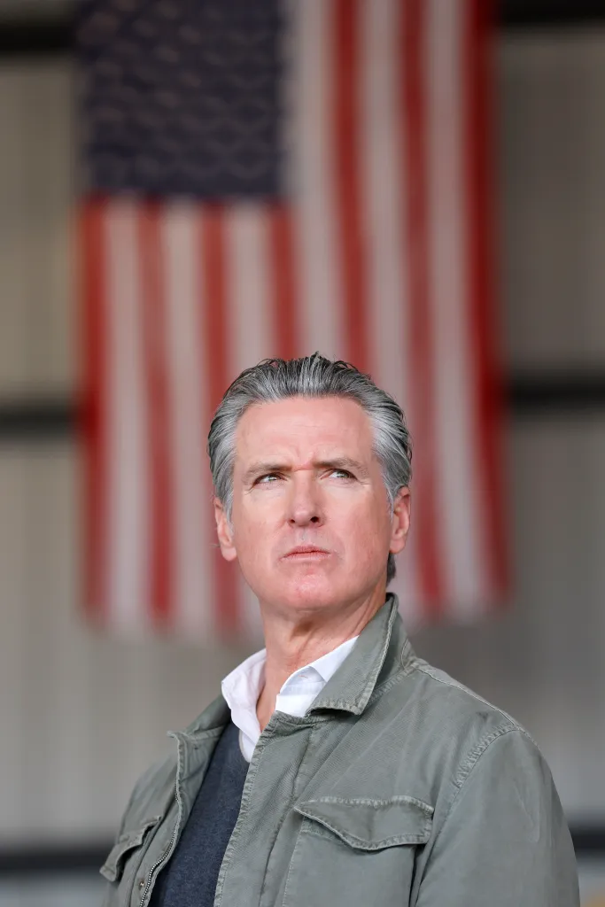 California Governor Gavin Newsom looking up and to the right during a news conference, with a blurred American flag in the background.