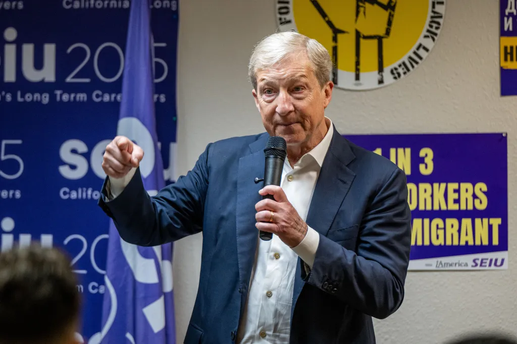 Tom Steyer speaking into a microphone, gesturing with his right hand, with a SEIU 2015 flag and a 