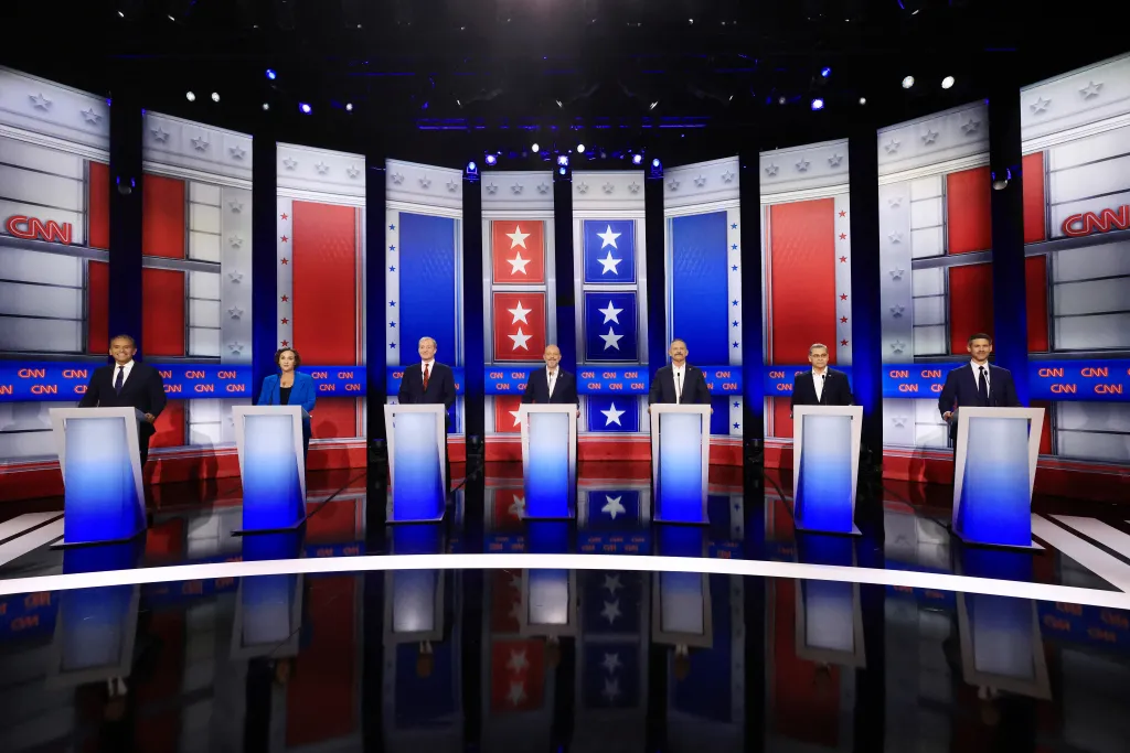 Seven candidates, Antonio Villaraigosa, Katie Porter, Tom Steyer, Steve Hilton, Chad Bianco, Xavier Becerra, and Matt Mahan, stand behind podiums on a CNN debate stage.