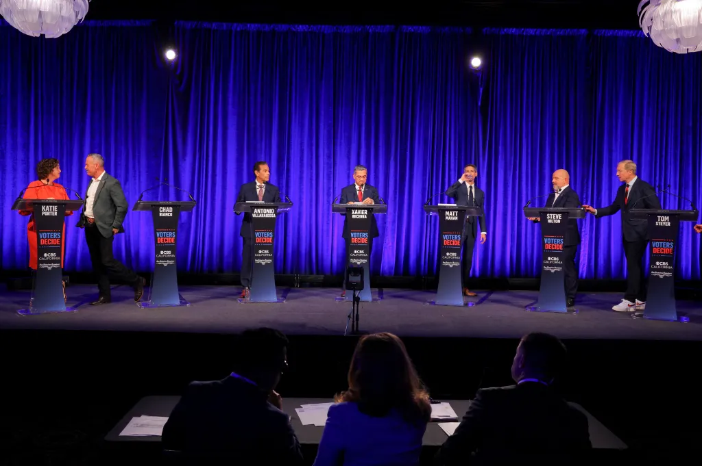 Candidates Katie Porter, Chad Bianco, Antonio Villaraigosa, Xavier Becerra, Matt Mahan, Steve Hilton, and Tom Steyer stand at podiums during a break at a California gubernatorial debate.