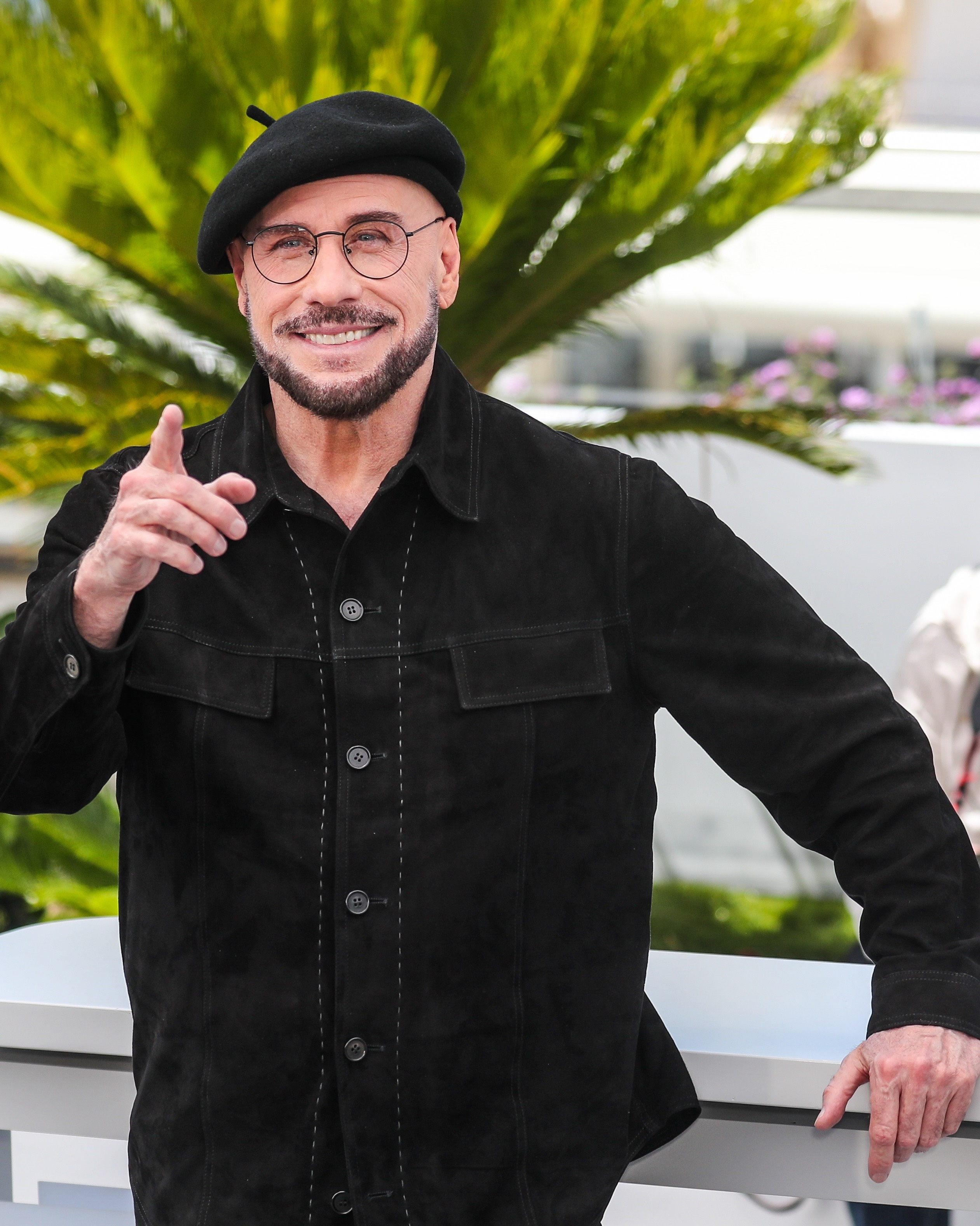 Actor John Travolta in a white beret, light pink tie, and black suit, pointing upwards at the 79th Cannes Film Festival.