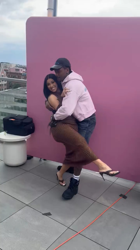 Cardi B and Stefon Diggs embrace each other on a rooftop against a pink wall.