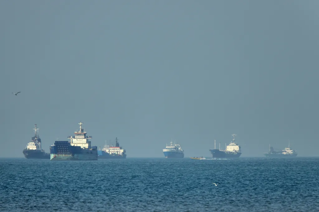 Cargo ships anchored in the Strait of Hormuz, with a small motorboat passing in the foreground.