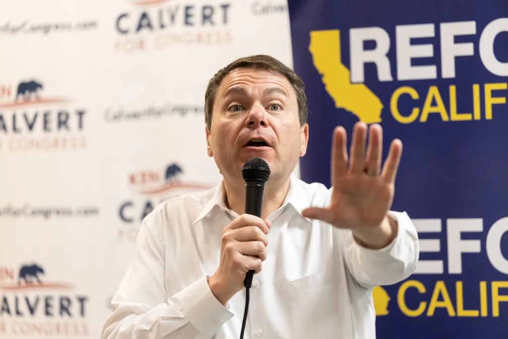 Carl DeMaio, California State Representative, speaking into a microphone with his left hand and gesturing with his right hand in front of a political banner.