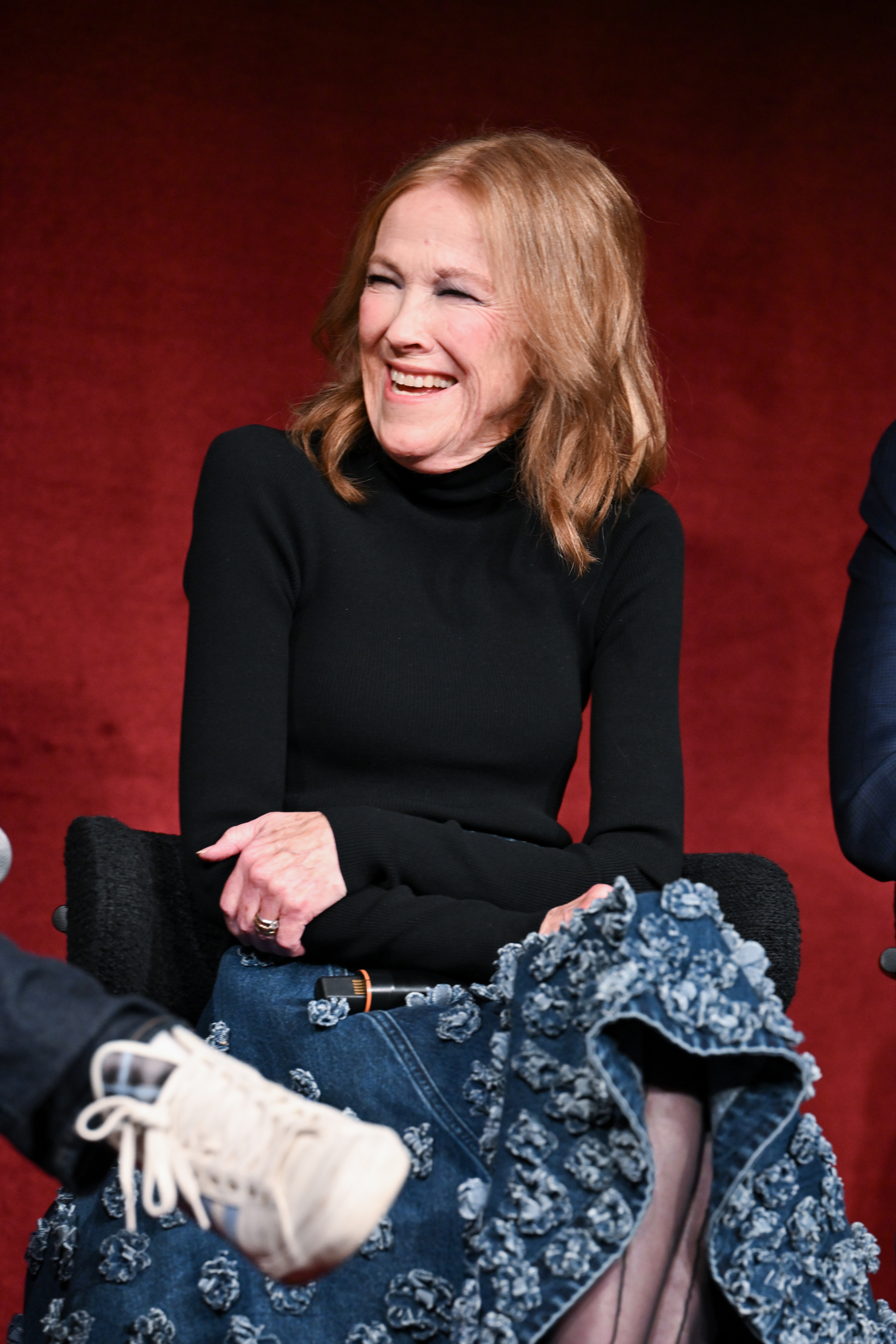 Catherine O'Hara smiles as she sits in a black turtleneck and an embellished denim skirt.