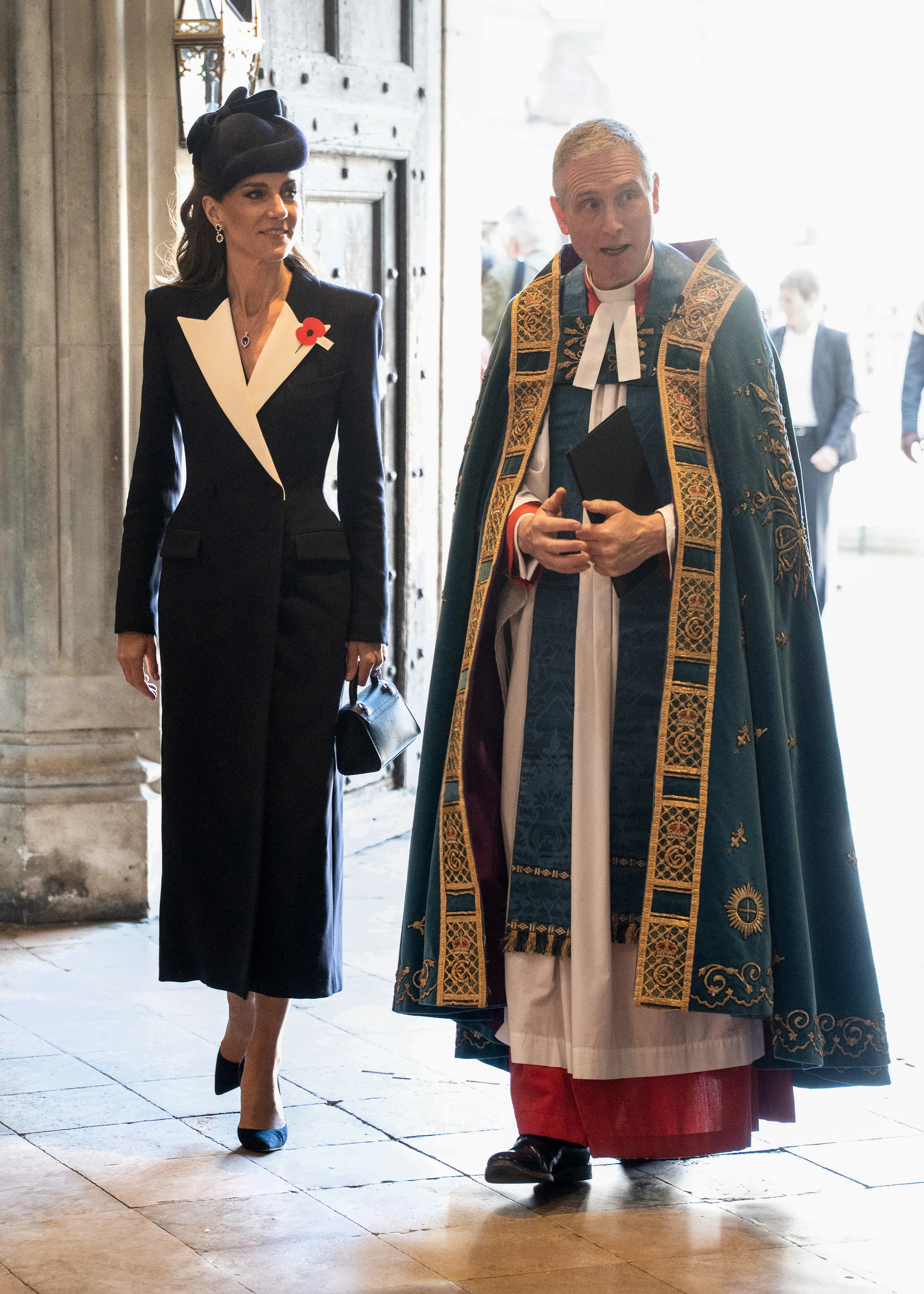 Catherine, Princess of Wales, in a black coat dress with a white lapel and a poppy pinned to it, walks with a robed clergyman at Westminster Abbey during an ANZAC Day service.