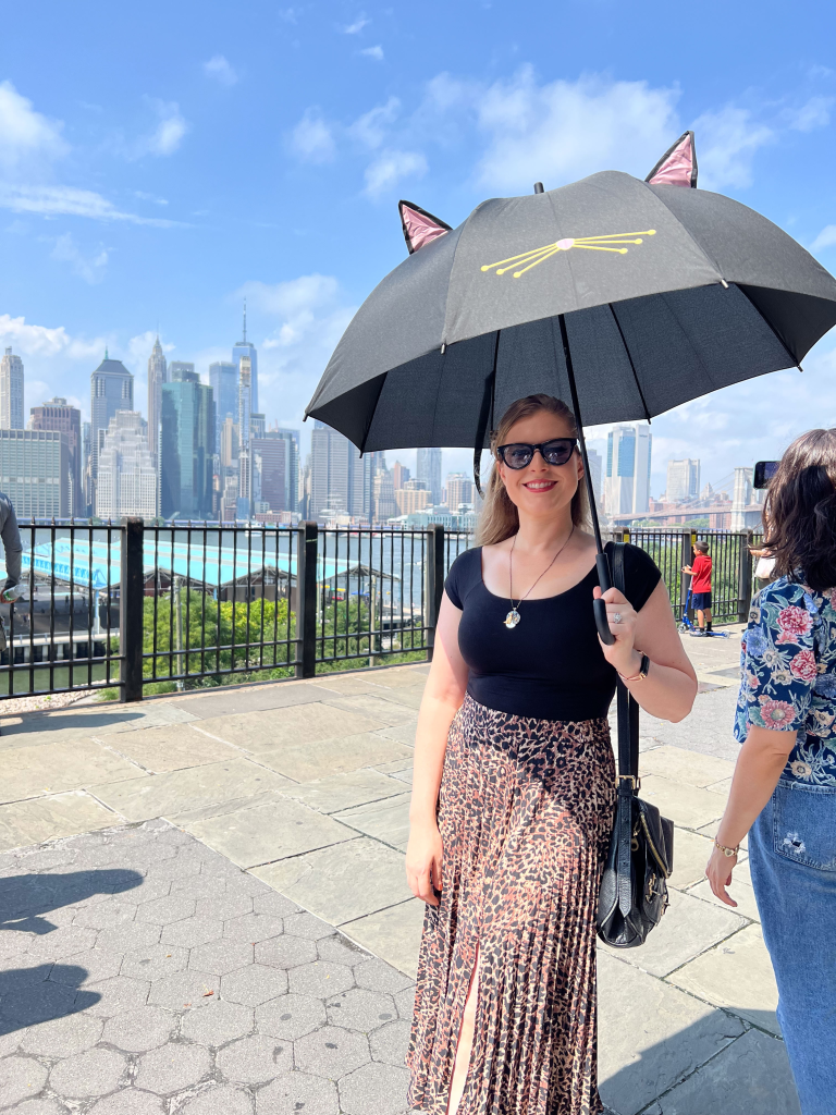 A woman holds a black umbrella with cat ears and whiskers as she smiles at the camera, with the New York City skyline visible behind her.