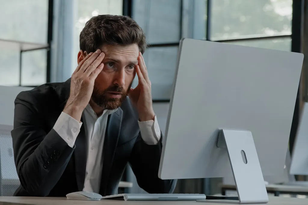 A stressed businessman clutches his head in front of a computer screen, frustrated by an online error or lost information.