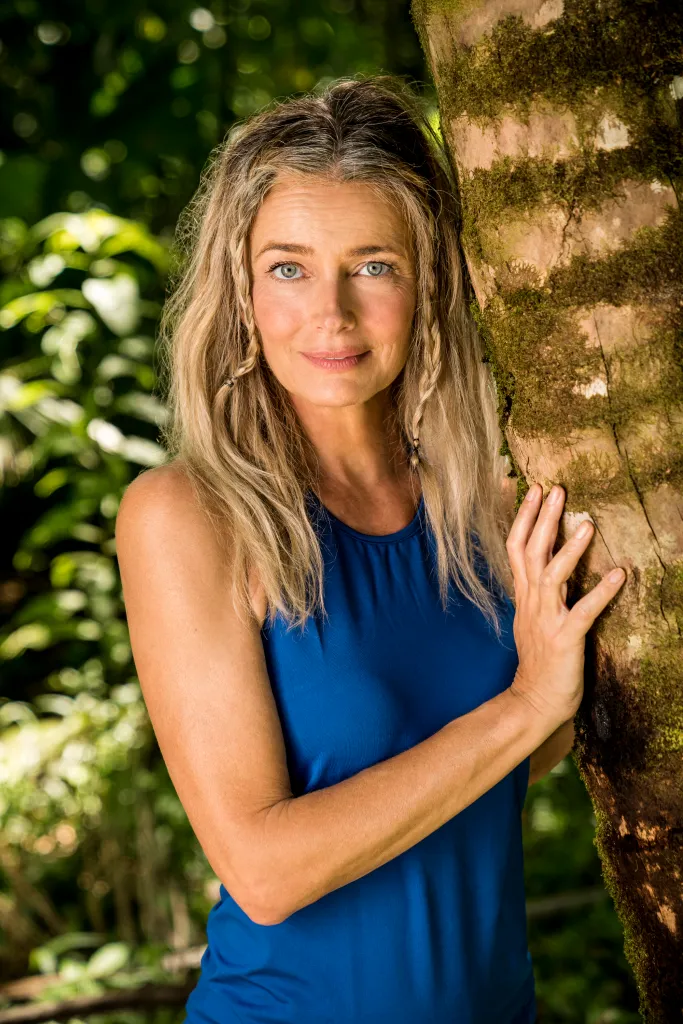 Paulina Porizkova in a blue top leans against a moss-covered tree.