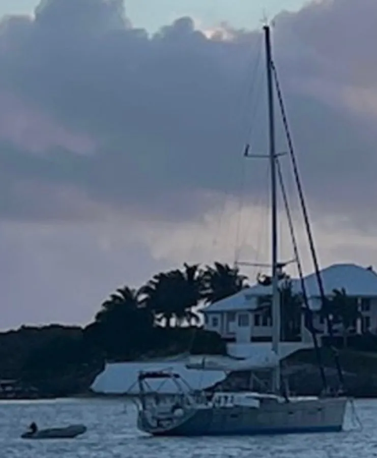 Sailboat anchored in water near land with houses and palm trees.