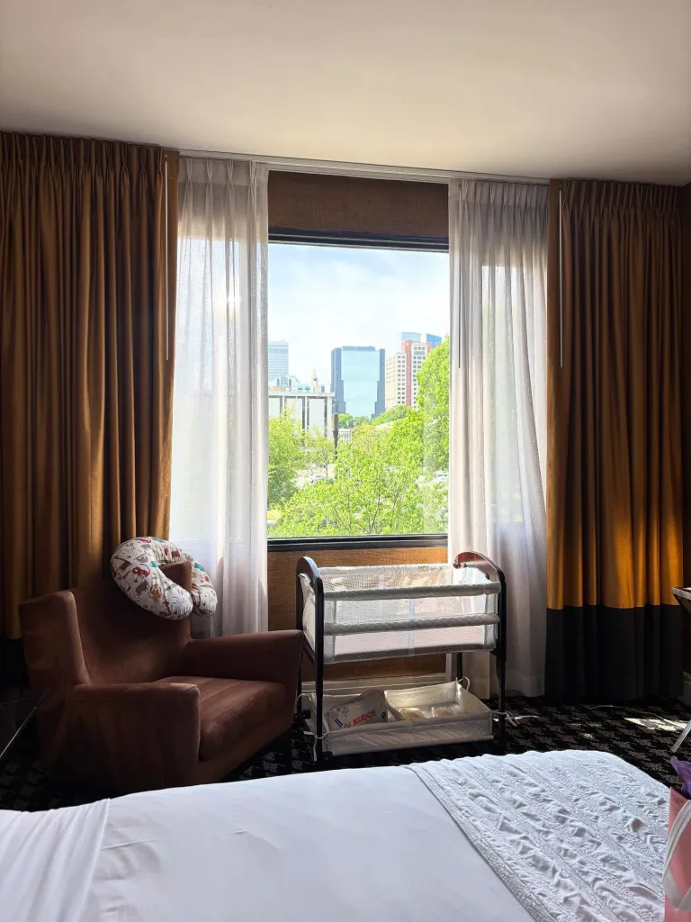 A hotel room in Charlotte, North Carolina with a bassinet and a chair with a nursing pillow next to a large window overlooking the city.