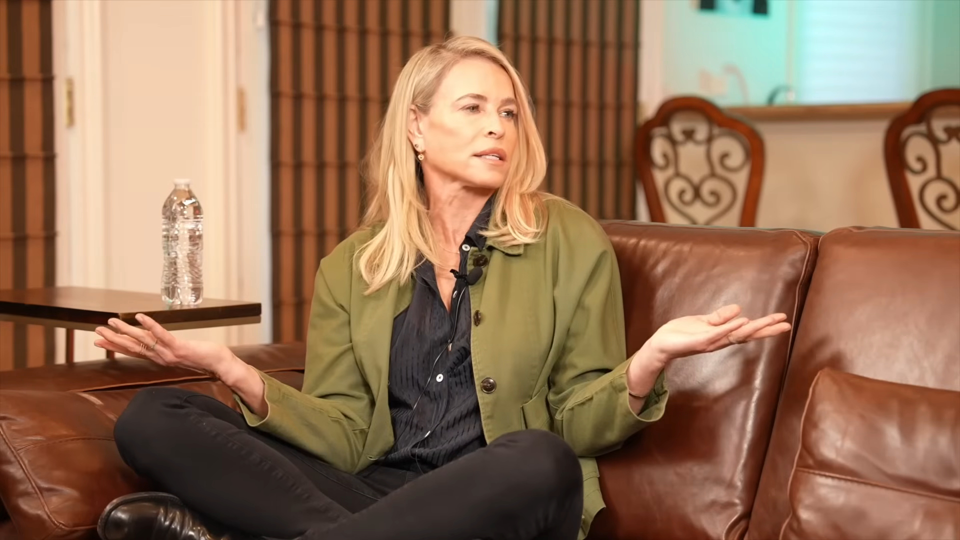 Chelsea Handler sitting on a brown leather couch, speaking with her hands outstretched.