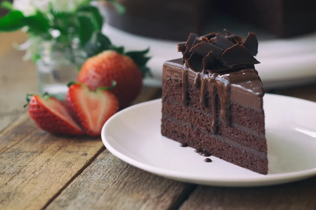 A slice of chocolate fudge cake with chocolate curls and a chocolate drizzle on a white plate, with strawberries and flowers in the background.